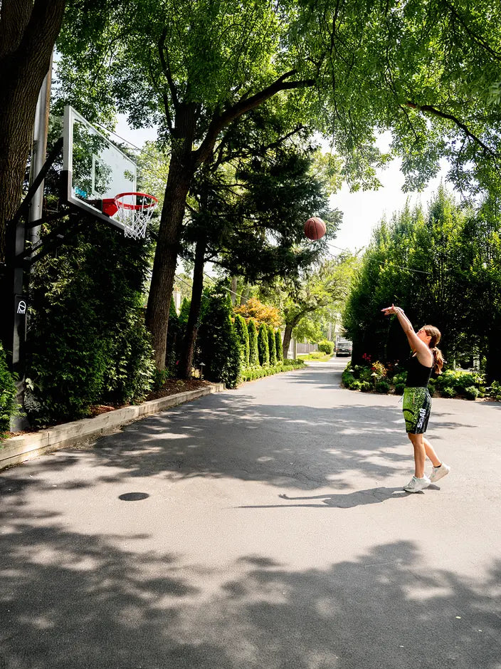 A daughter practicing her jump shot at a SOAR hoop on a tree-lined driveway